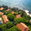 Aerial shot of red-roofed villas by a clear blue sea, surrounded by lush greenery.