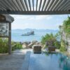 Elegant wooden deck with infinity pool overlooking serene seaside view with mountains and boat.