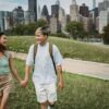 Positive diverse couple in casual apparel holding hands while walking on grass lawn near contemporary buildings and looking at each other